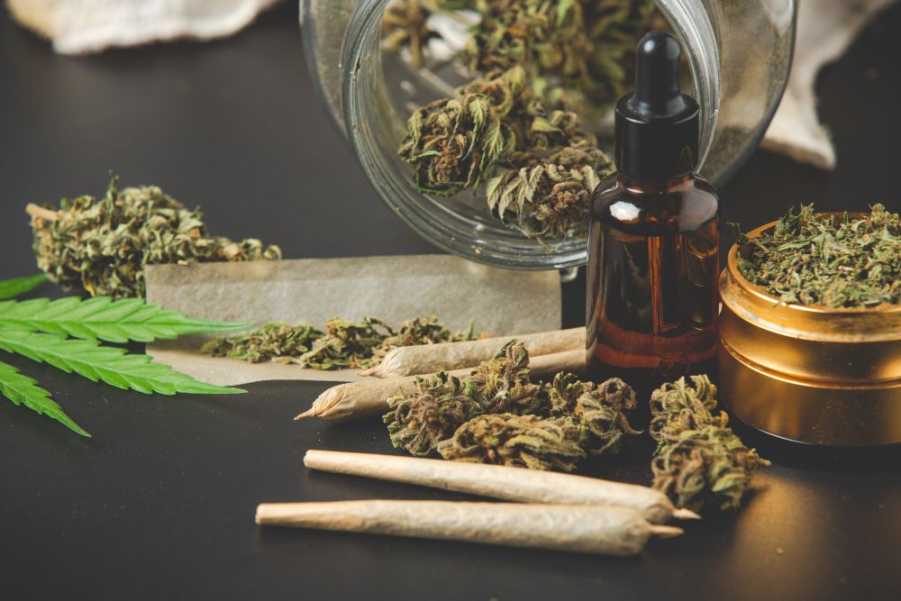 Close-up of cannabis buds, pre-rolled joints, hemp leaf, jar, grinder and CBD oil bottle on a dark surface. 