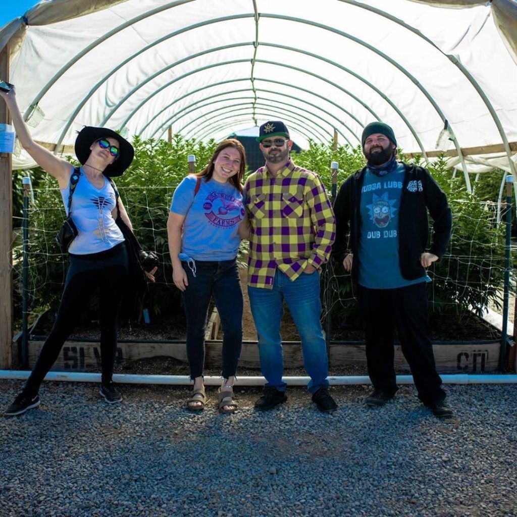 Four people stand inside a white greenhouse with plants visible in the background.