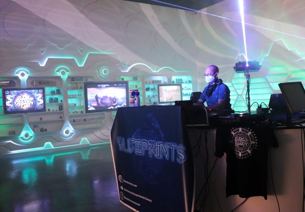 A DJ wearing a mask performs in a futuristic-looking room with neon lights. A banner reads