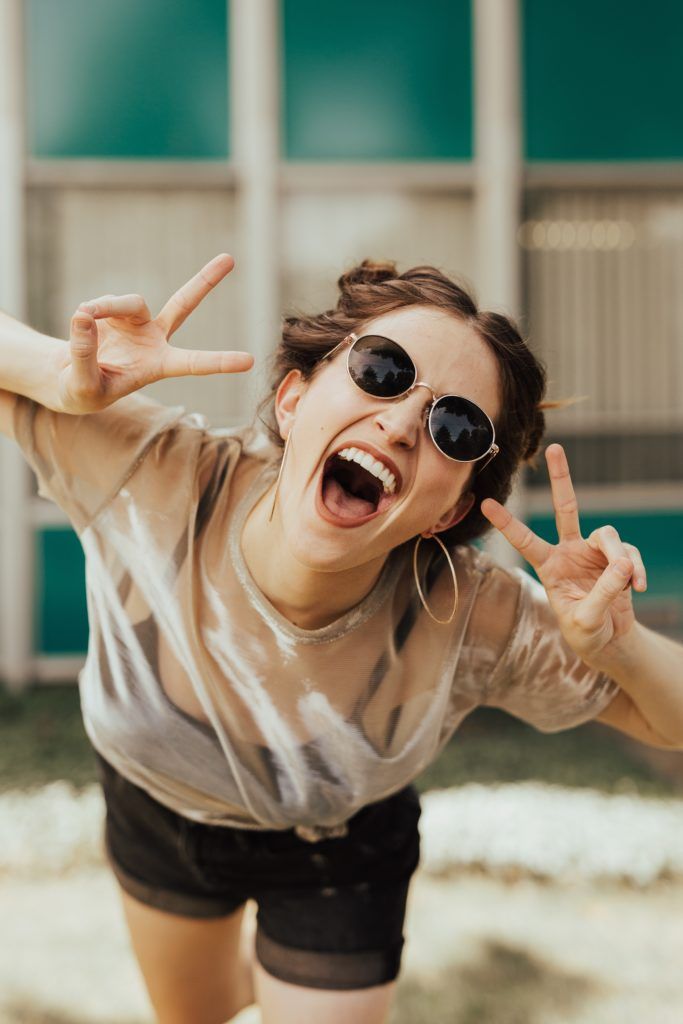 Woman wearing sunglasses makes peace signs, mouth open wide in an excited expression, outside a building.