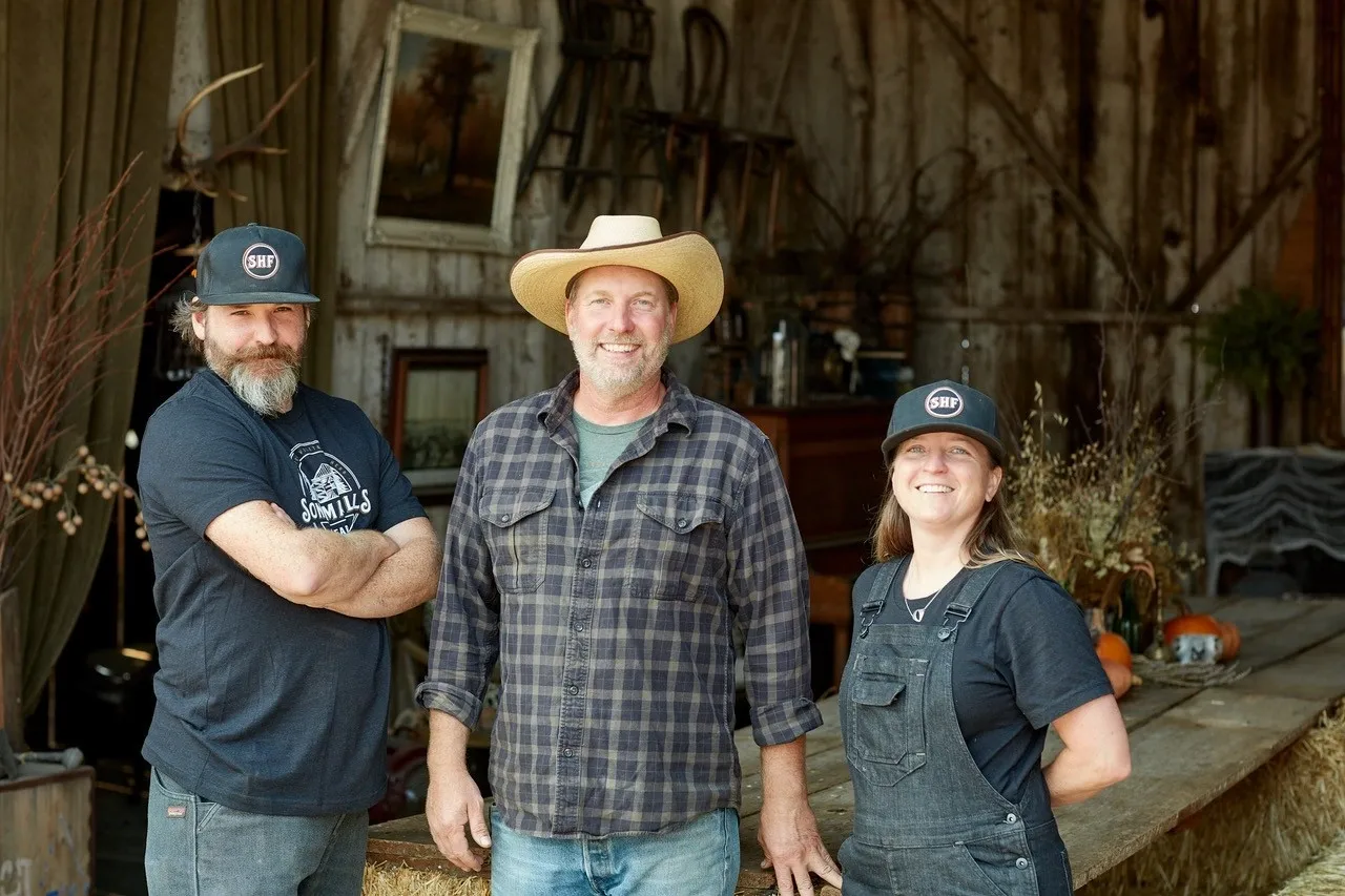 Three people in work clothes standing in a rustic barn, with pumpkins and farm tools in the background.