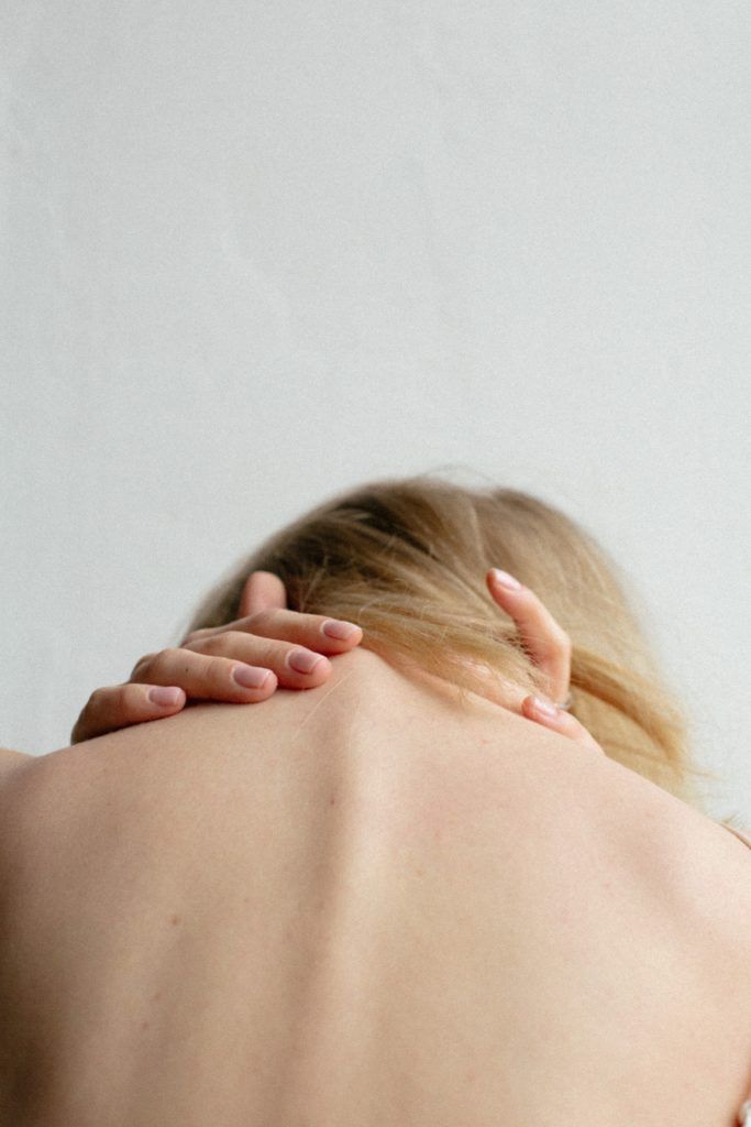 Woman with blonde hair, seen from the back, holding her neck, likely in pain, against a plain white background.