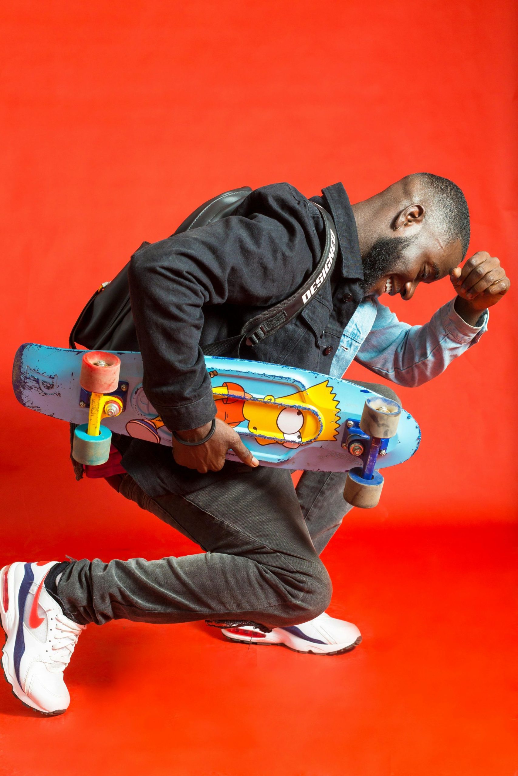 Man in blue and black holds a skateboard with a Simpsons design against a red background, with an arm raised.